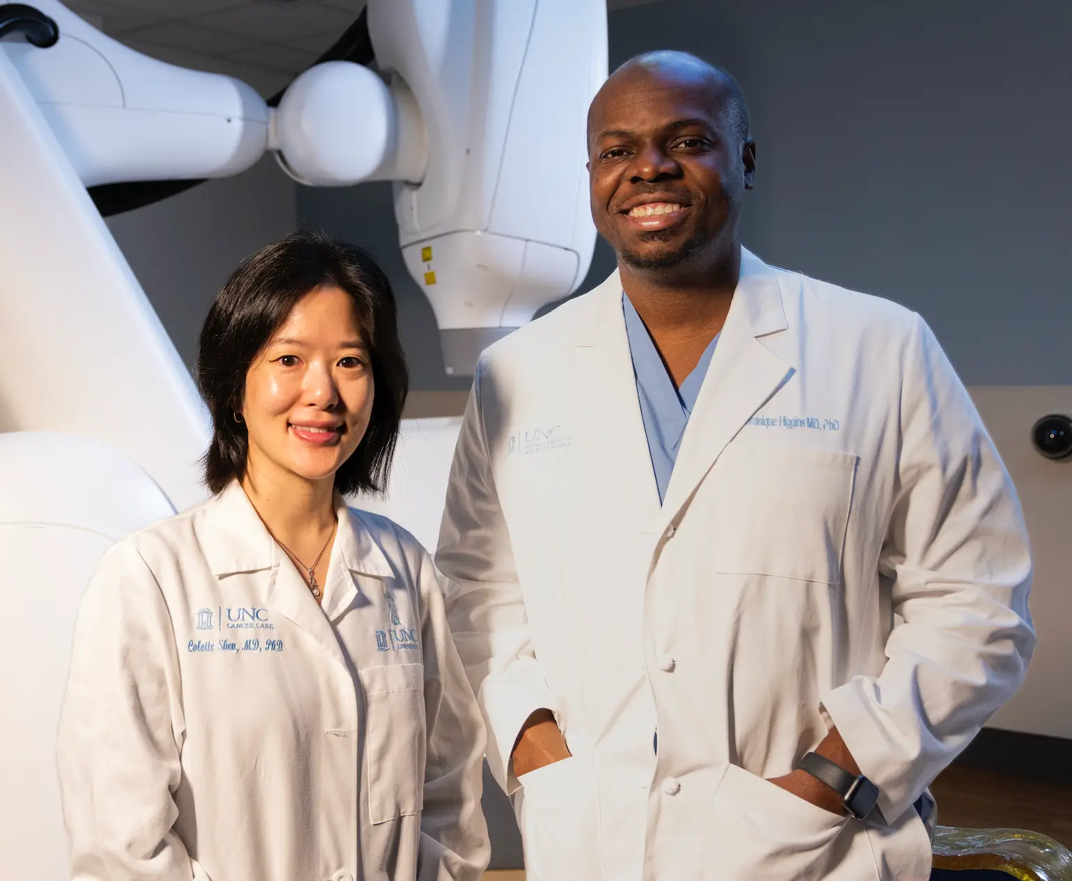 Two UNC Health cancer research specialists stand beside advanced radiation therapy equipment, supporting innovative cancer treatment and patient care made possible by research donors
