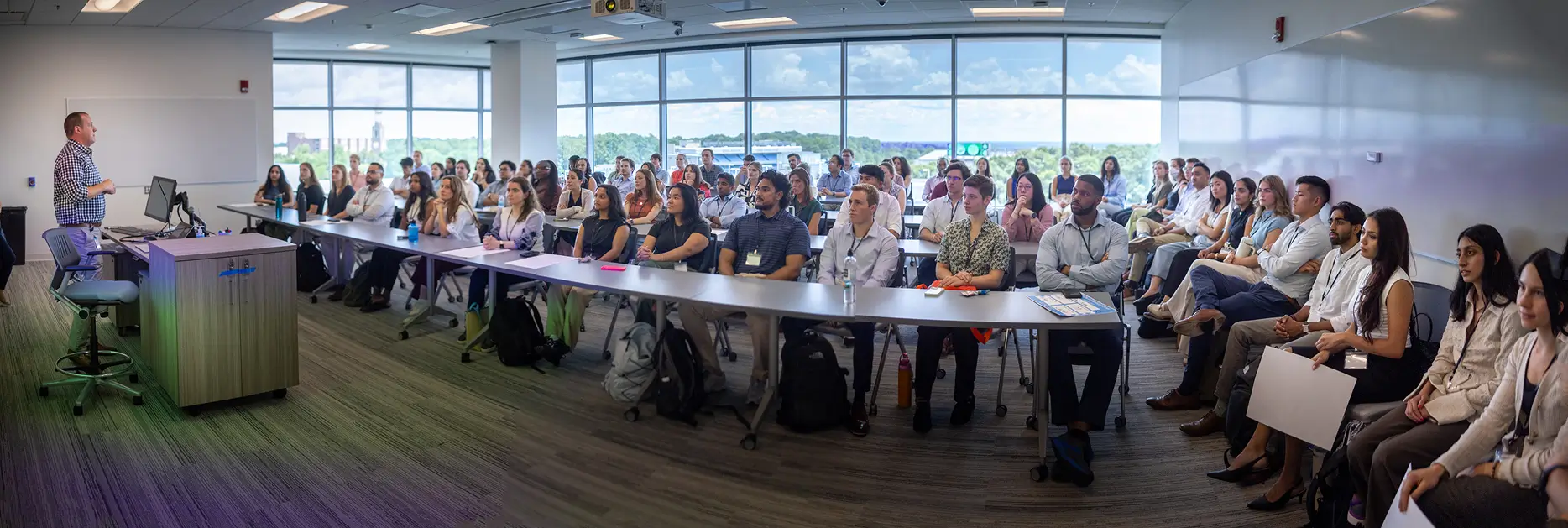 UNC School of Medicine research students attend a large orientation session, listening to a faculty presentation in a modern classroom overlooking the campus.