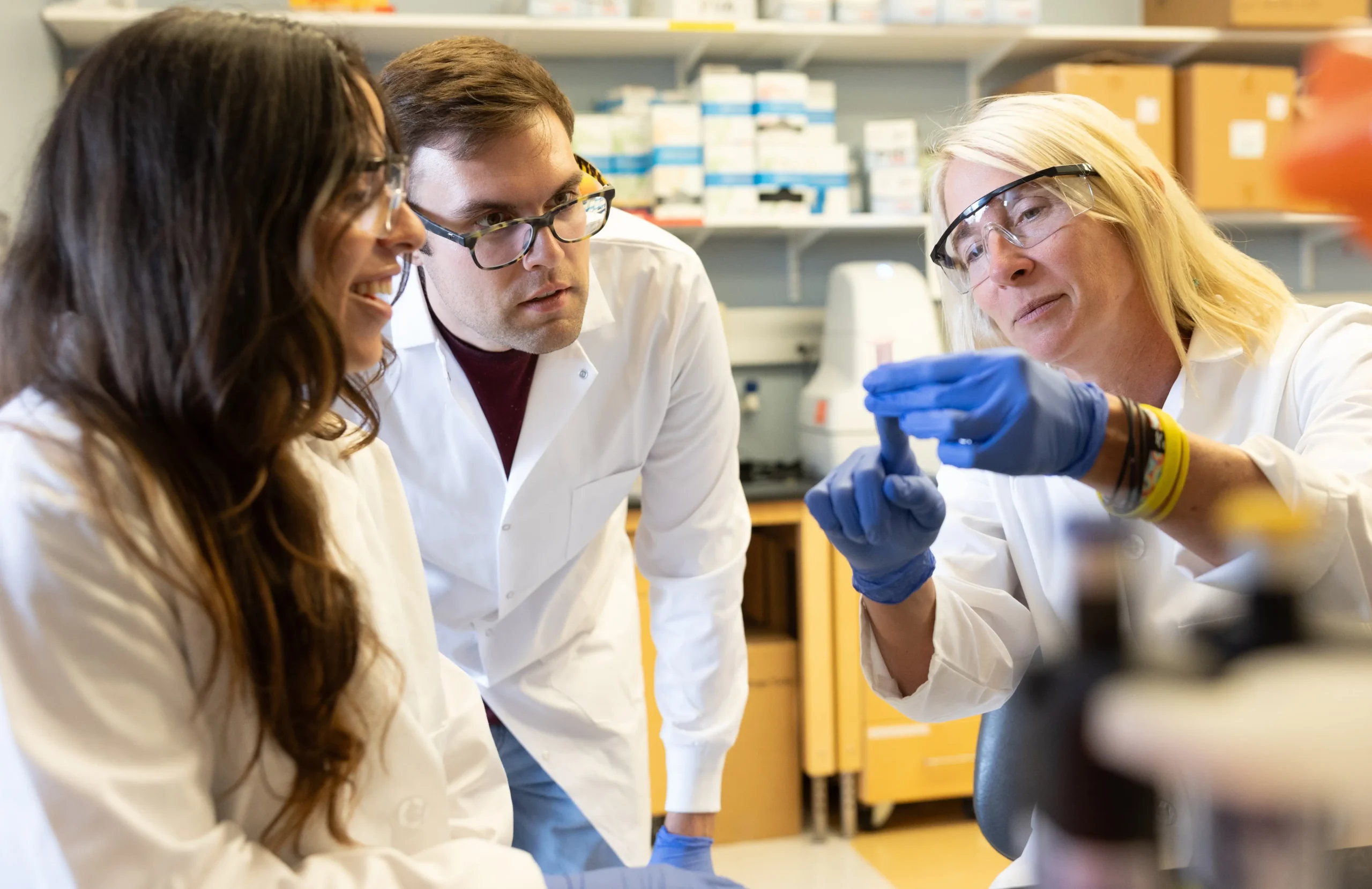 Three medical researchers collaborate in a cancer research laboratory to examine a sample using gloved hands.