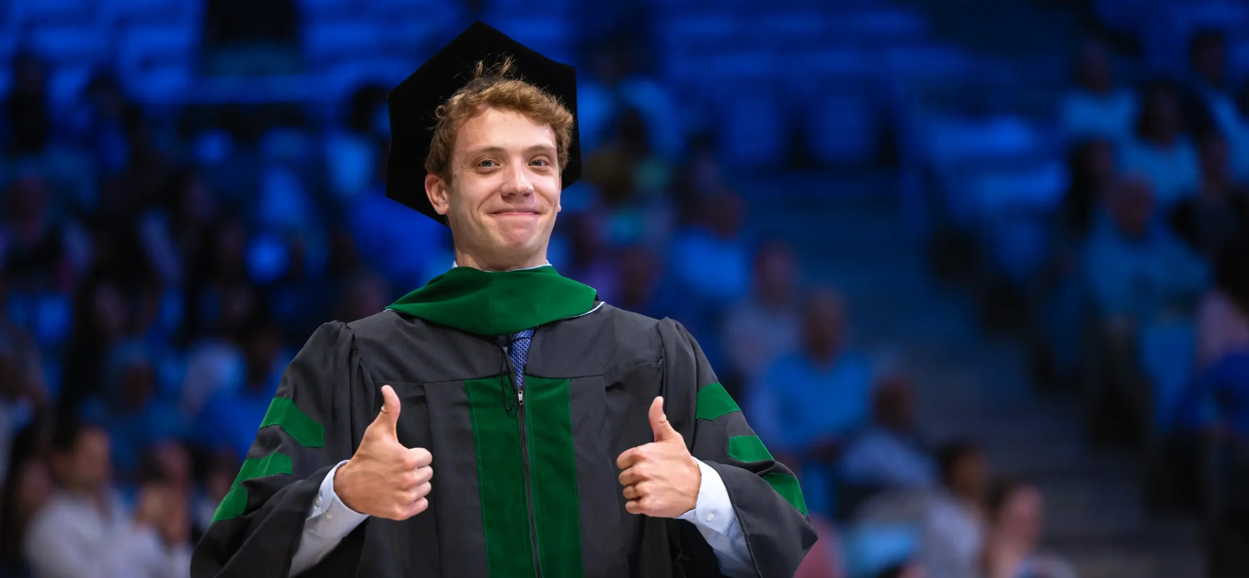 UNC medical graduate in a black and green doctoral gown and cap gives two thumbs up during a graduation ceremony, completing an education made possible by scholarship donations