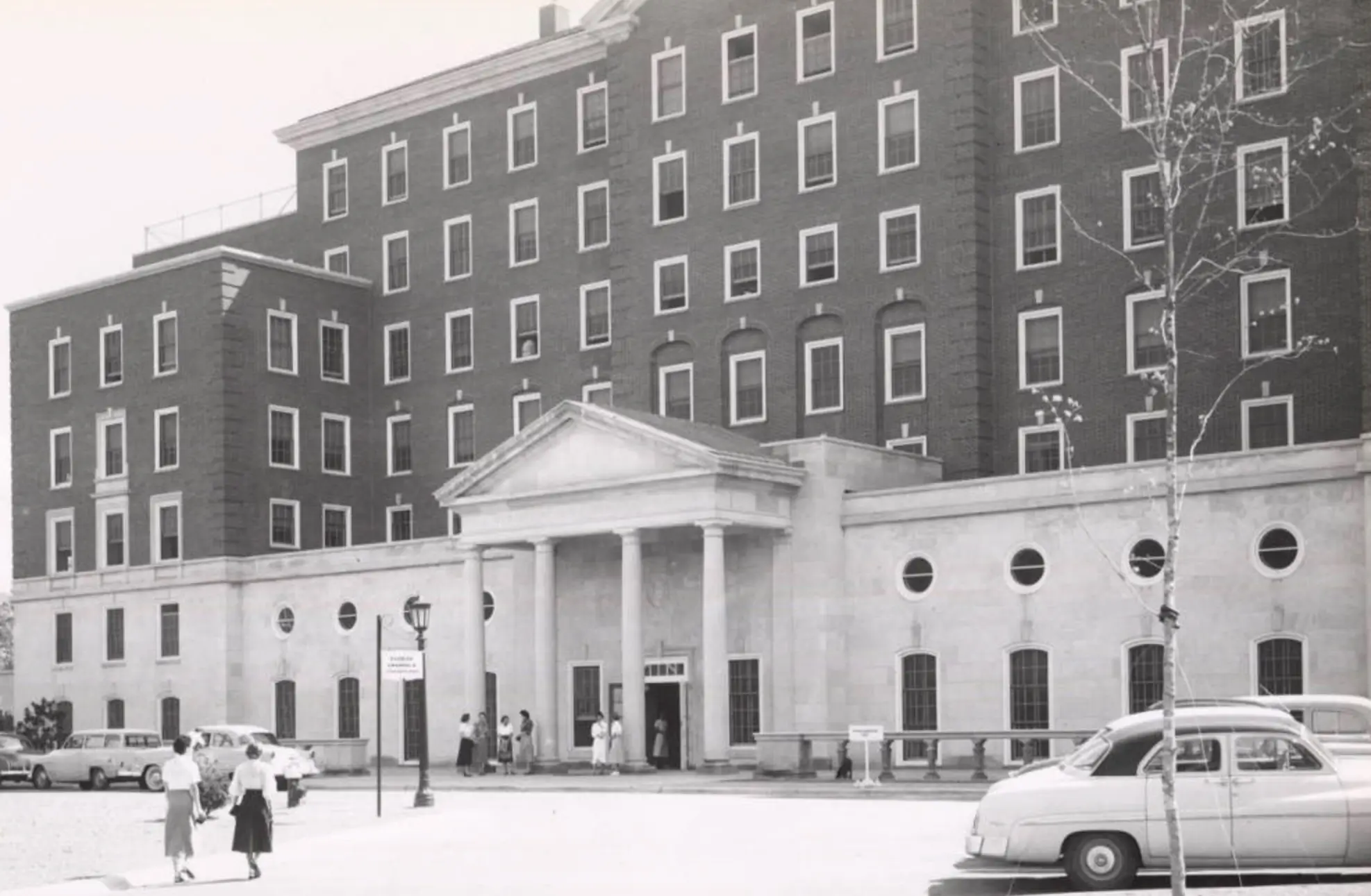 Historic black-and-white photograph of a UNC research hospital building with classic architecture, vintage automobiles, and visitors gathered outside the main entrance.