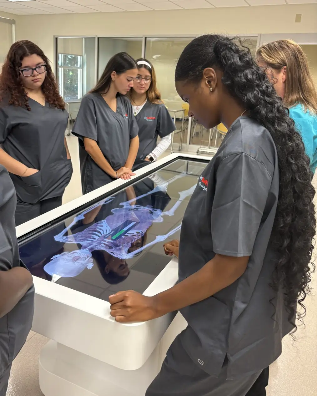Medical students in scrubs use a virtual anatomy dissection table to explore a 3D human skeleton during an interactive learning session at UNC funded by healthcare philanthropy