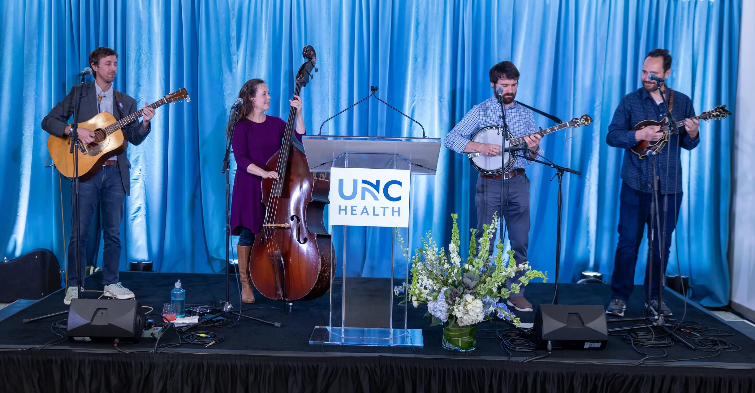 Musicians perform live on stage at a UNC Health event, featuring guitar, upright bass, banjo, and mandolin.