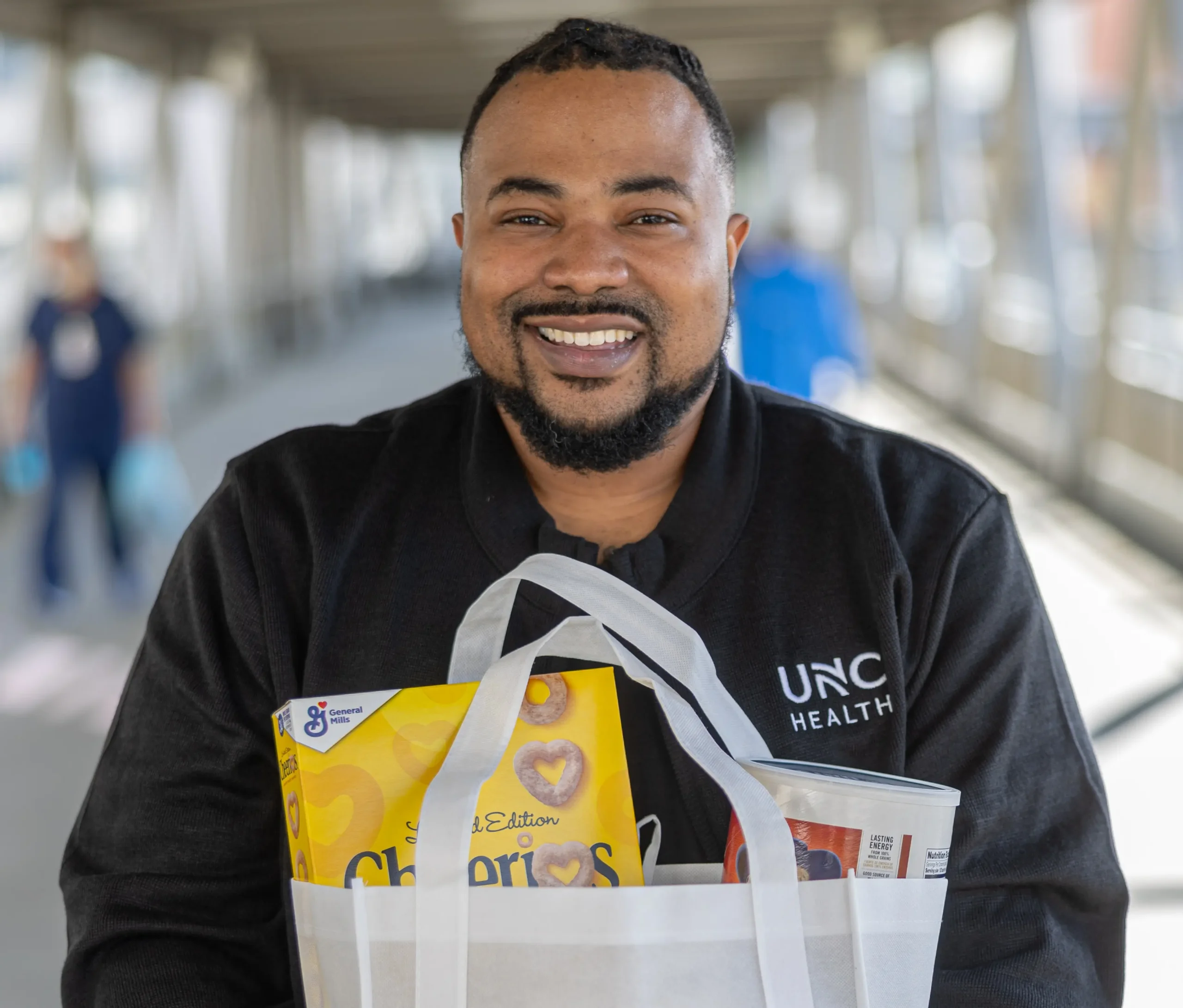 Smiling UNC Health team member holding a bag of donated food for patients in need in the hospital pedestrian walkway.