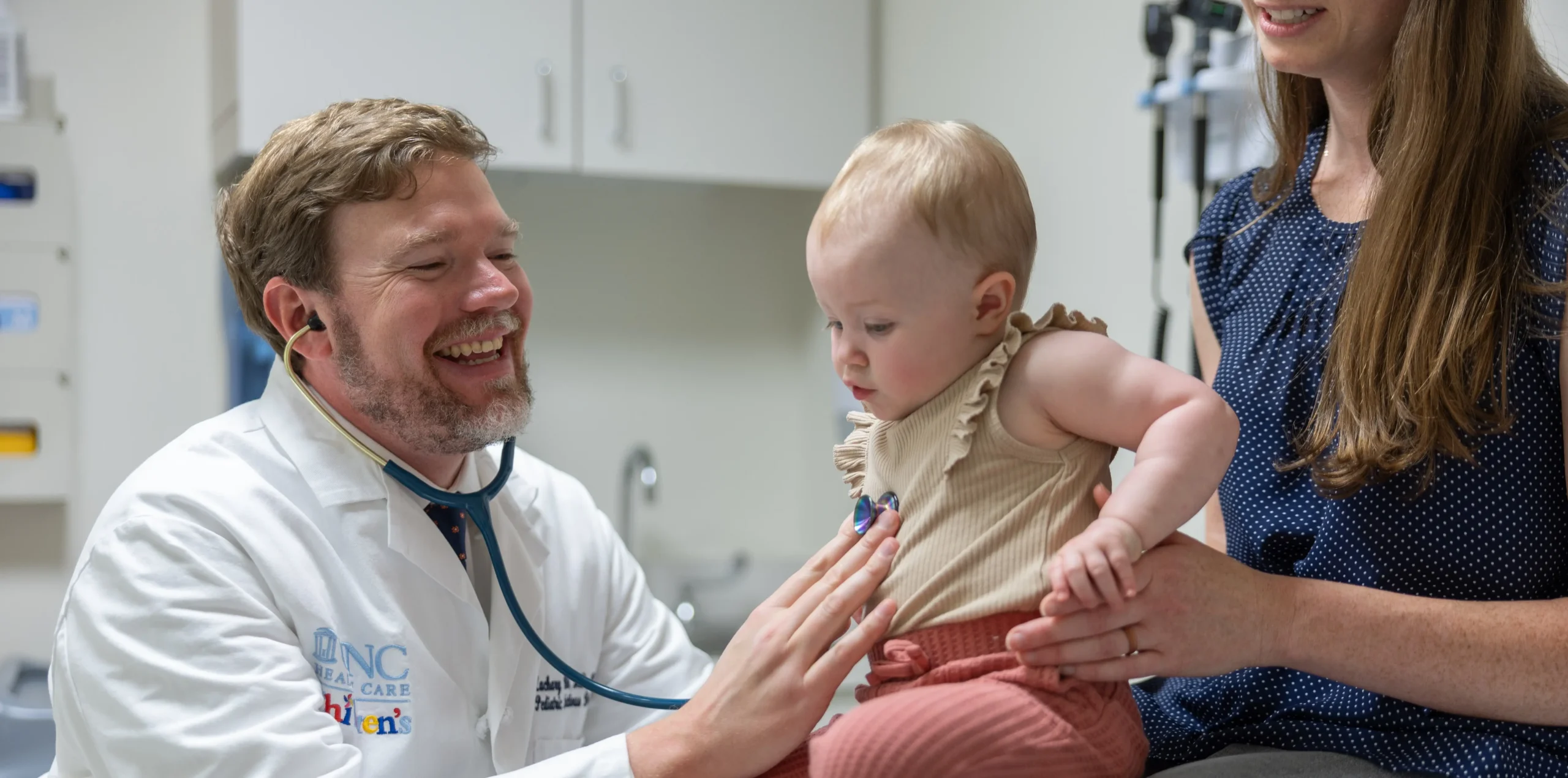 UNC Children’s pediatrician uses a stethoscope while examining a baby during a routine wellness visit as a parent looks on in the exam room.