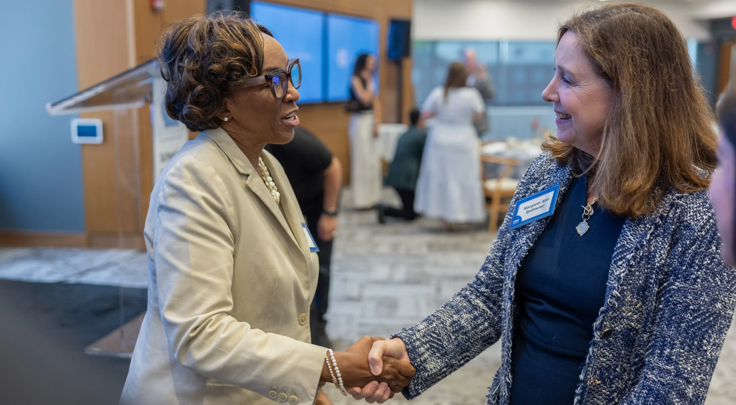 Two professionals smiling and shaking hands during a leadership or staff event in a modern indoor setting.
