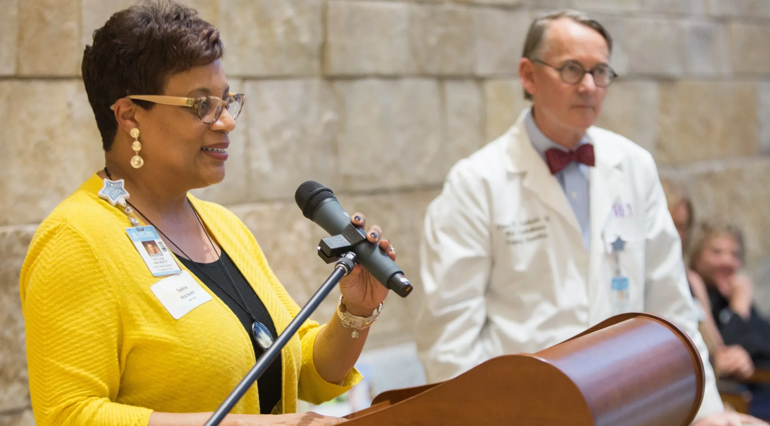 Healthcare professional speaking at a podium during a community event, with a physician standing nearby.