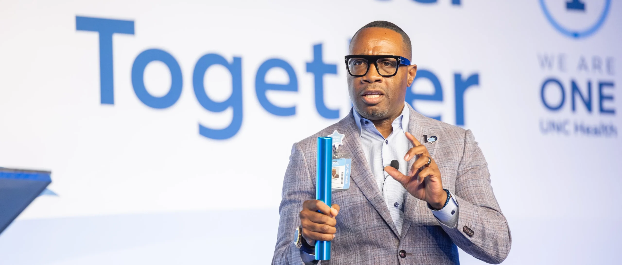 UNC Health leader speaks on stage during a staff event, delivering a presentation on how foundation grants facilitate statewide service for patients in rural areas of NC.