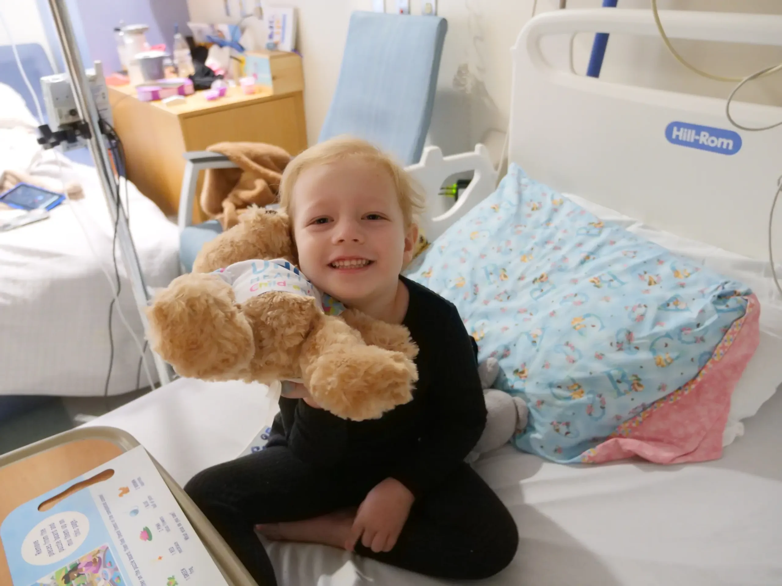 Smiling child sitting in a hospital bed and holding a teddy bear, benefitting from foundation grants for cancer research