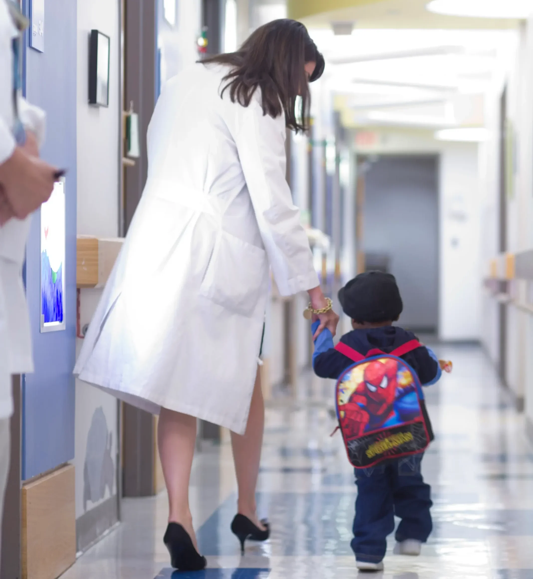 UNC Children’s cancer researcher walks hand in hand with a young patient wearing a Spider-Man backpack through a pediatric hospital hallway.