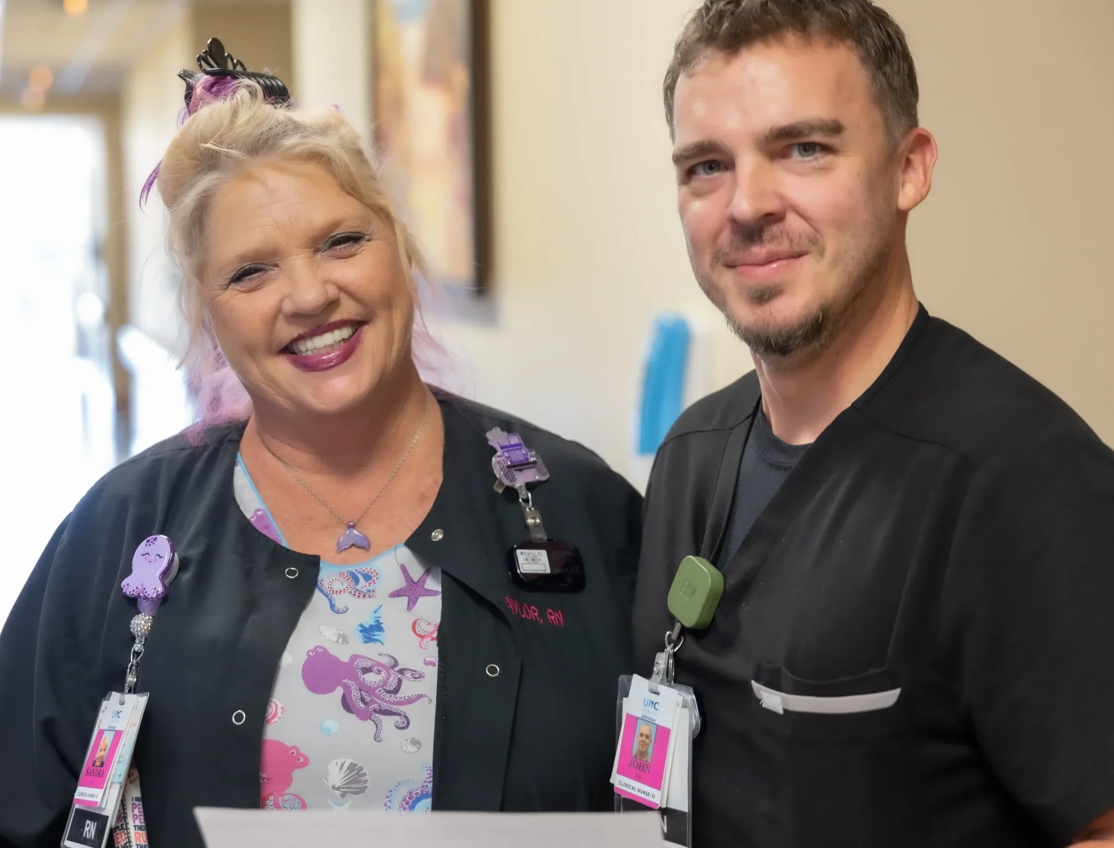 Two smiling healthcare workers wearing medical uniforms and employee ID badges in a hospital setting.