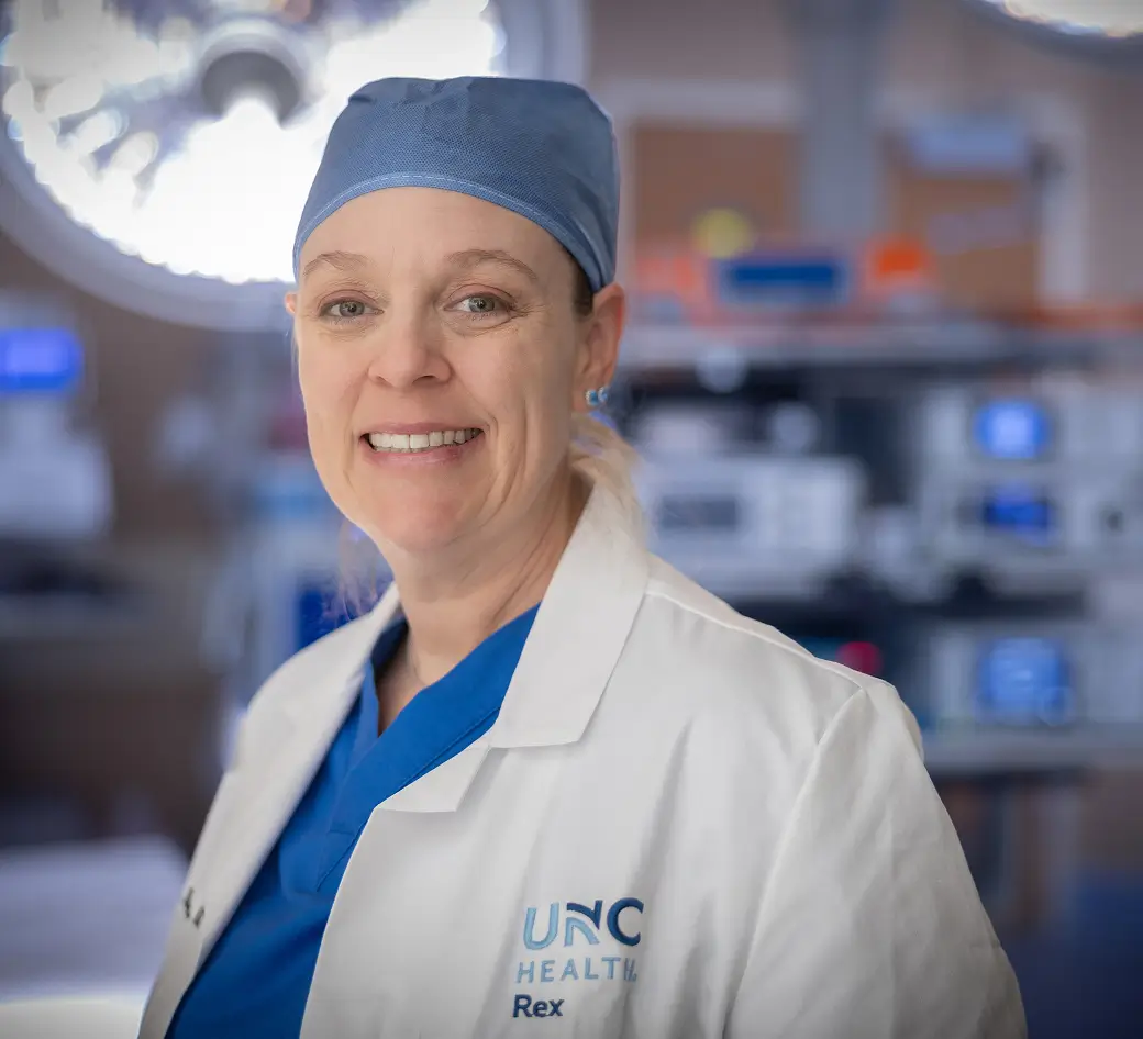 Smiling surgeon wearing surgical scrubs, cap, and a white medical coat standing inside a modern operating room supported through foundation funds.