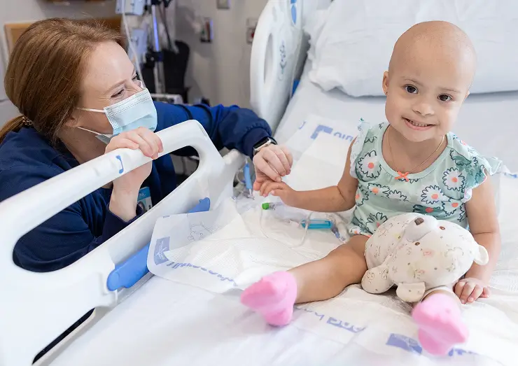 Young pediatric cancer patient smiling in a hospital bed while holding hands with a childhood cancer researcher
