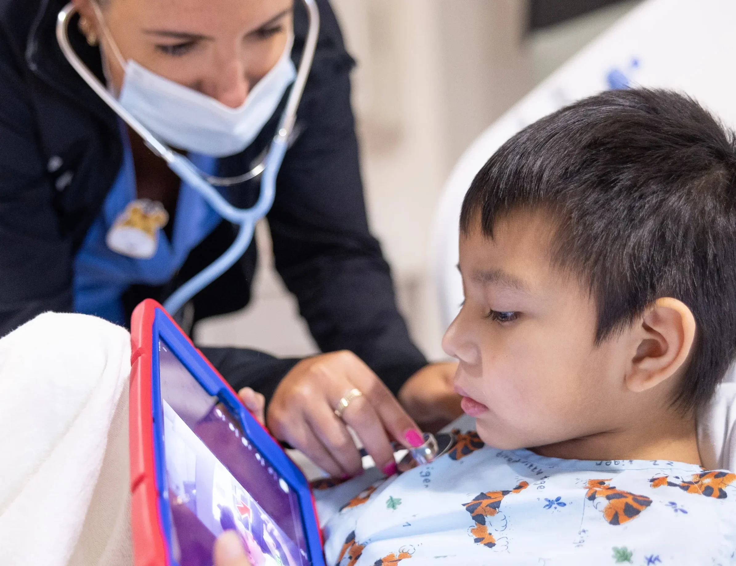 UNC Children’s healthcare provider listens to a young patient’s chest with a stethoscope while the child watches a tablet in a hospital room.