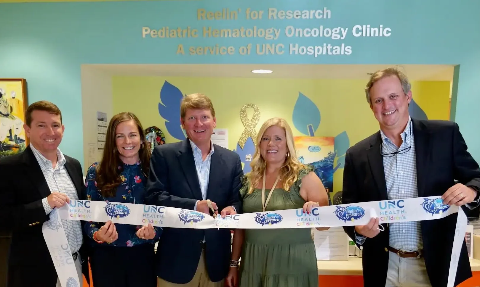 Five people holding a Reelin’ for Research and UNC Health Children’s ribbon at a pediatric hematology oncology clinic opening.