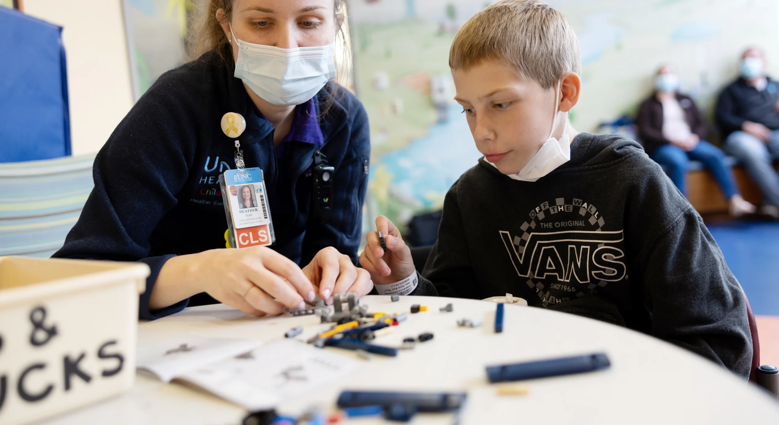 Pediatric healthcare staff member helping a child with a hands-on activity at a table in a hospital setting.